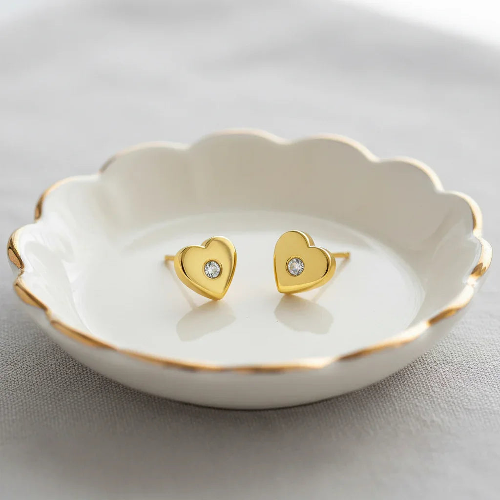 A pair of gold earrings with a heart-shaped accent sits on a white, scalloped-edge plate.